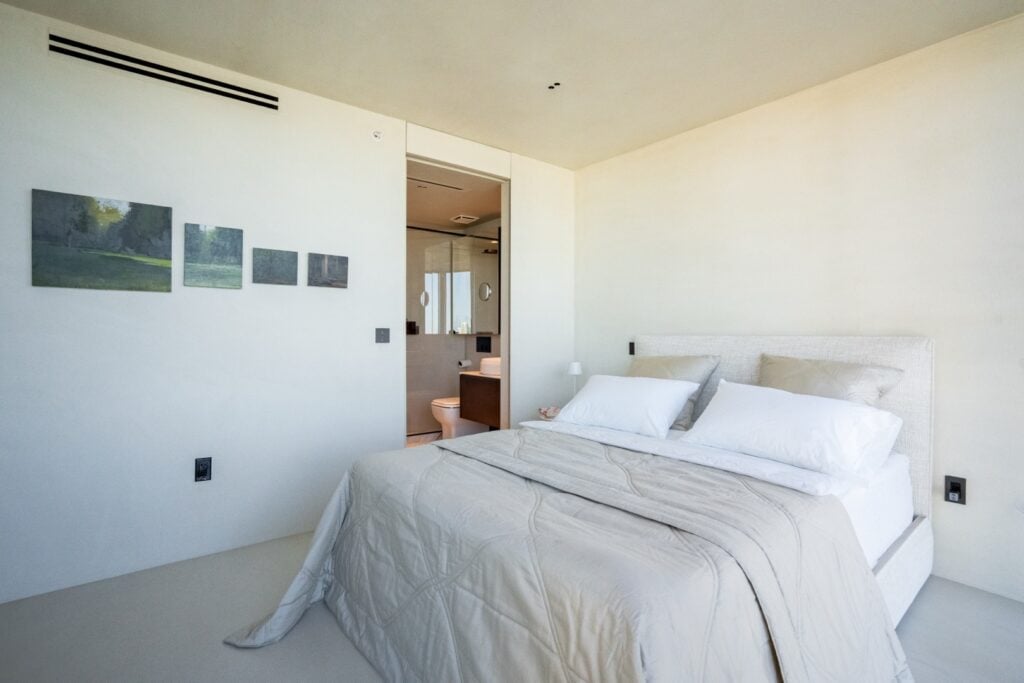 A minimalist bedroom with a neatly made bed, beige bedding, wall art, and an open door leading to a modern bathroom.