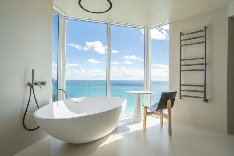 Modern bathroom with a freestanding oval bathtub, a black chair, a small white table, towel warmer, and large windows overlooking the ocean on a sunny day.
