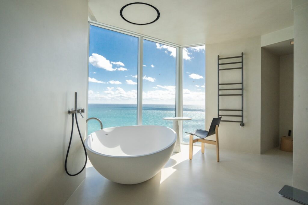 Modern bathroom with a freestanding oval tub, floor-mounted faucet, minimalist chair, towel rack, and large window overlooking the ocean under a blue sky with clouds.