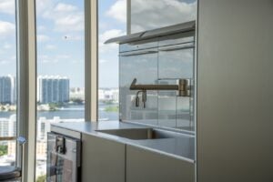 A modern kitchen sink with a metallic faucet, reflective backsplash, and a window view of high-rise buildings and water in the background.