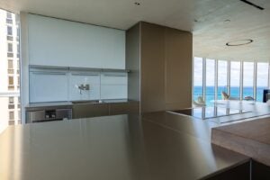 Modern kitchen with minimalist design and metallic surfaces, featuring large windows with a view of the ocean and a building exterior visible on the left.