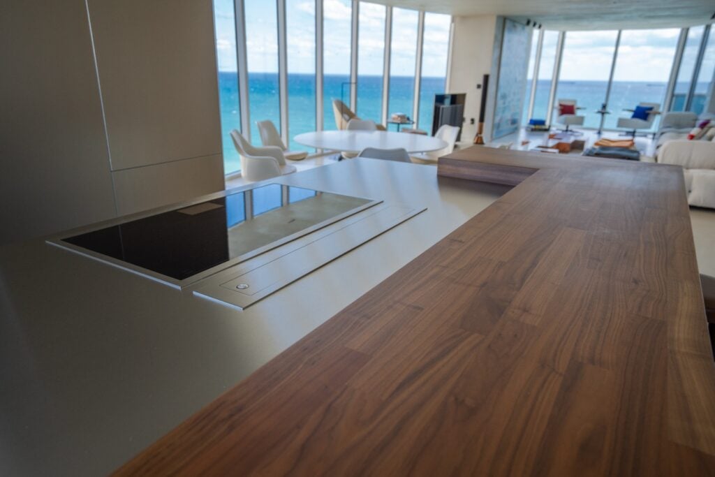 Modern open-plan kitchen with a sleek cooktop and wooden countertop, overlooking a dining and living area with floor-to-ceiling windows and an ocean view.