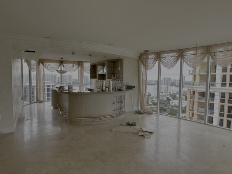 An empty high-rise apartment with marble floors, a built-in bar, large windows with city views, and sheer drapes; some clutter and debris are on the floor.